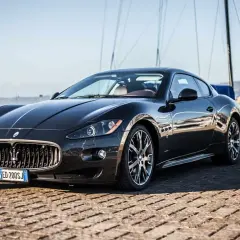 Eleganckie, czarne coupe auto Maserati stoi na nabrzeżu, z żaglami łodzi w tle.