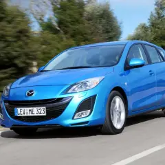 Niebieska Mazda 3 2010 w wersji hatchback pędzi po krętej drodze wśród zieleni.