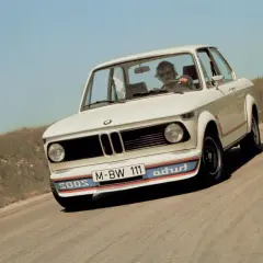 Białe BMW E20 z kolorowymi pasami na zderzaku i czerwonymi akcentami na karoserii jedzie po krętej drodze.