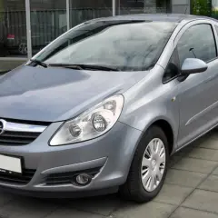 Srebrny Opel Corsa D 1.4 benzyna stoi na chodniku przed budynkiem z dużymi oknami.