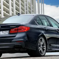 Ciemny BMW m550i z napędem xDrive, z widocznym logo BMW i oznaczeniem modelu, zaparkowany przed nowoczesnym budynkiem.