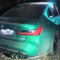 Zielony BMW M3 Competition, z napisem "bogus m3" na tylnej szybie, stoi nocą na leśnej drodze.