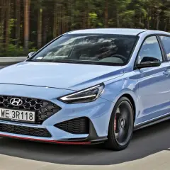 Błękitny Hyundai i30 N, sportowy hatchback, pędzi po drodze wśród drzew.