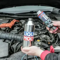 Dwie ręce trzymają produkty Liqui Moly do czyszczenia katalizatora. W tle widać silnik samochodu.