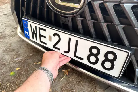 Dłoń poprawia polskie tablice rejestracyjne WE 2JL88 na grillu samochodu.