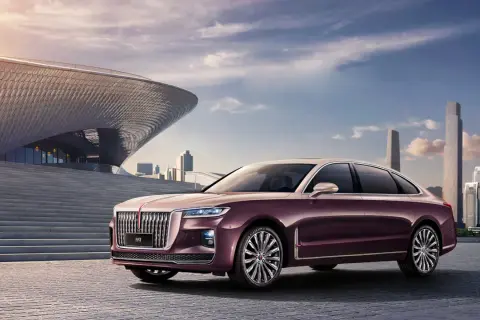 Elegancki, bordowy Hongqi H9 parkuje przed nowoczesnym budynkiem o falistej fasadzie, w tle miejska panorama.