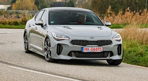 Szary Kia Stinger GT z kierowcą za kierownicą jedzie po asfaltowej drodze, otoczony suchą trawą i drzewami.