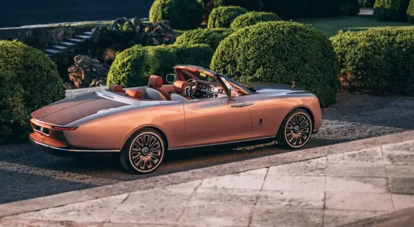 Elegancki Rolls-Royce Boat Tail w kolorze miedzianego różu, z drewnianym tyłem, zaparkowany wśród bujnej zieleni.