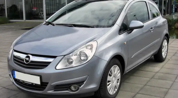 Srebrny Opel Corsa D 1.4 benzyna stoi na chodniku przed budynkiem z dużymi oknami.