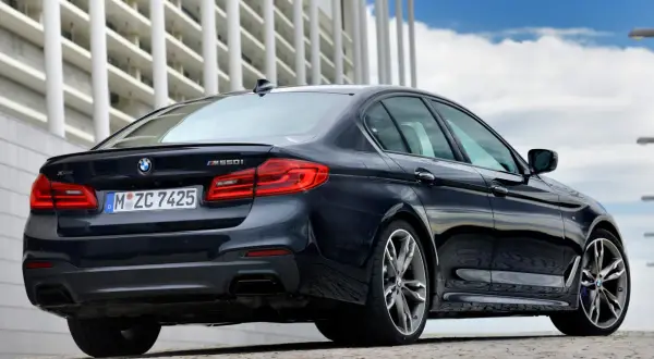 Ciemny BMW m550i z napędem xDrive, z widocznym logo BMW i oznaczeniem modelu, zaparkowany przed nowoczesnym budynkiem.