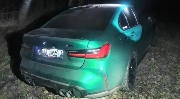 Zielony BMW M3 Competition, z napisem "bogus m3" na tylnej szybie, stoi nocą na leśnej drodze.