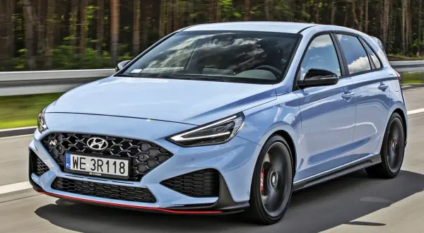 Błękitny Hyundai i30 N, sportowy hatchback, pędzi po drodze wśród drzew.