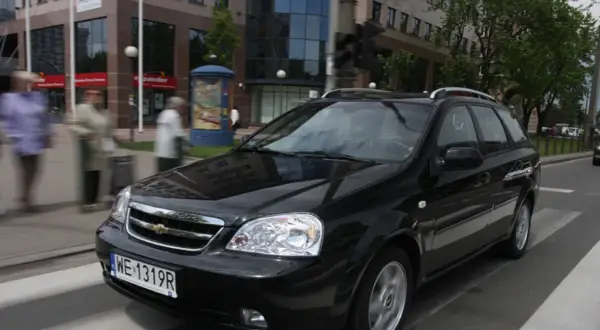 Czarny Chevrolet Lacetti kombi jedzie ulicą, mijając budynki i przechodniów.