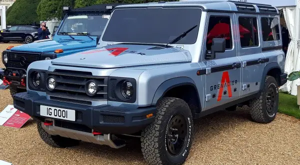 Srebrny INEOS Grenadier z czerwonym logo "A" na drzwiach, gotowy na przygodę. Obok niebieski SUV.