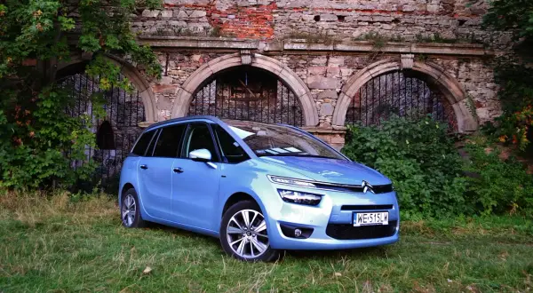 Błękitny Citroen C4 Grand Picasso 2.0 HDI zaparkowany na trawie przed zabytkowym murem z cegły i kamienia.