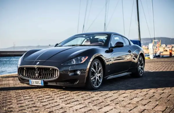 Eleganckie, czarne coupe auto Maserati stoi na nabrzeżu, z żaglami łodzi w tle.