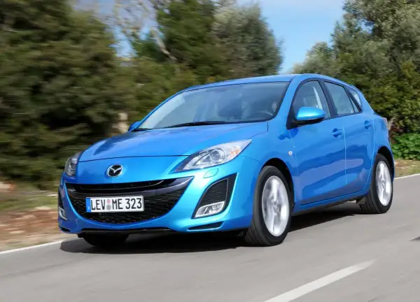 Niebieska Mazda 3 2010 w wersji hatchback pędzi po krętej drodze wśród zieleni.