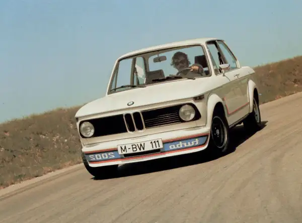 Białe BMW E20 z kolorowymi pasami na zderzaku i czerwonymi akcentami na karoserii jedzie po krętej drodze.