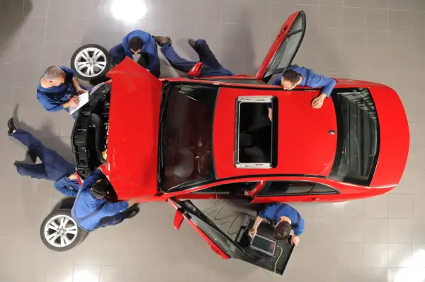 Mechanicy pracują przy czerwonym aucie. Jeden spisuje notatki, inny podłącza komputer diagnostyczny. Wszyscy skupieni, by zapobiec deprecjacji pojazdu.
