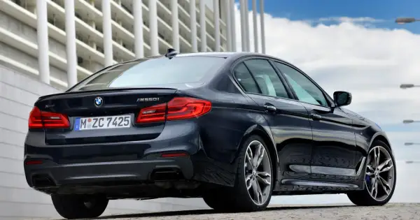 Ciemny BMW m550i z napędem xDrive, z widocznym logo BMW i oznaczeniem modelu, zaparkowany przed nowoczesnym budynkiem.