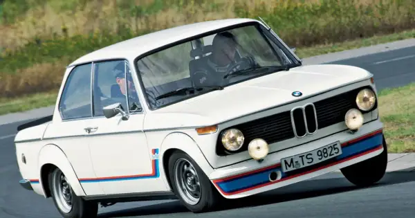 Białe BMW 2002 Turbo z kolorowymi pasami na zderzaku i błotnikach, pędzi po torze.