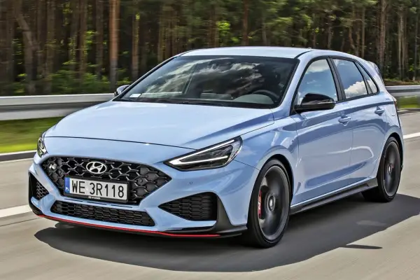 Błękitny Hyundai i30 N, sportowy hatchback, pędzi po drodze wśród drzew.