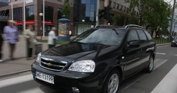 Czarny Chevrolet Lacetti kombi jedzie ulicą, mijając budynki i przechodniów.
